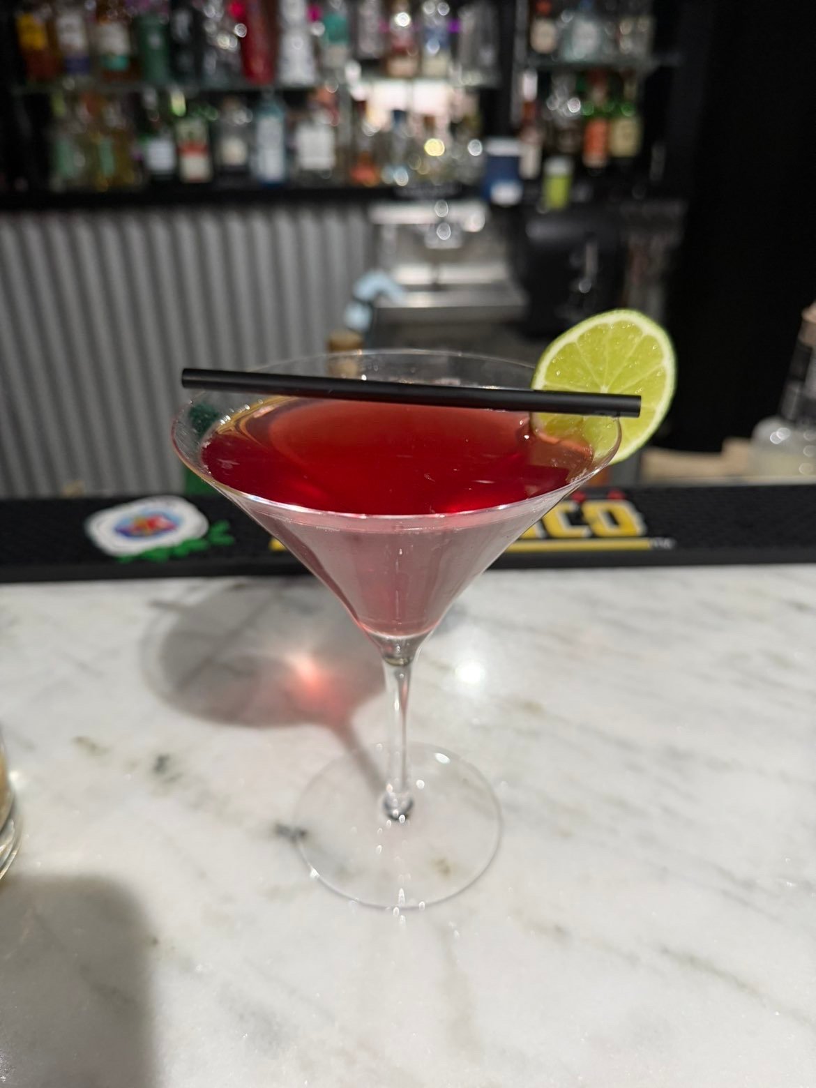 Red cocktail in a martini glass with lime wheel garnish and black straw on marble bar counter