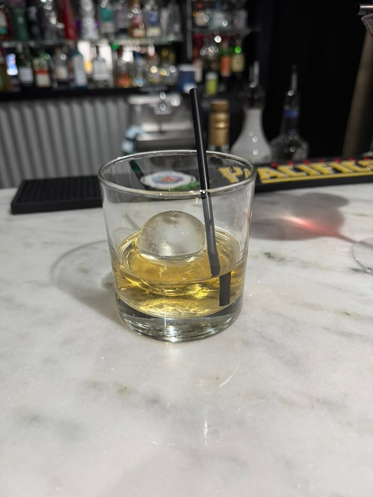 Whiskey glass with ice and black straw on marble bar counter with blurred bottles in background