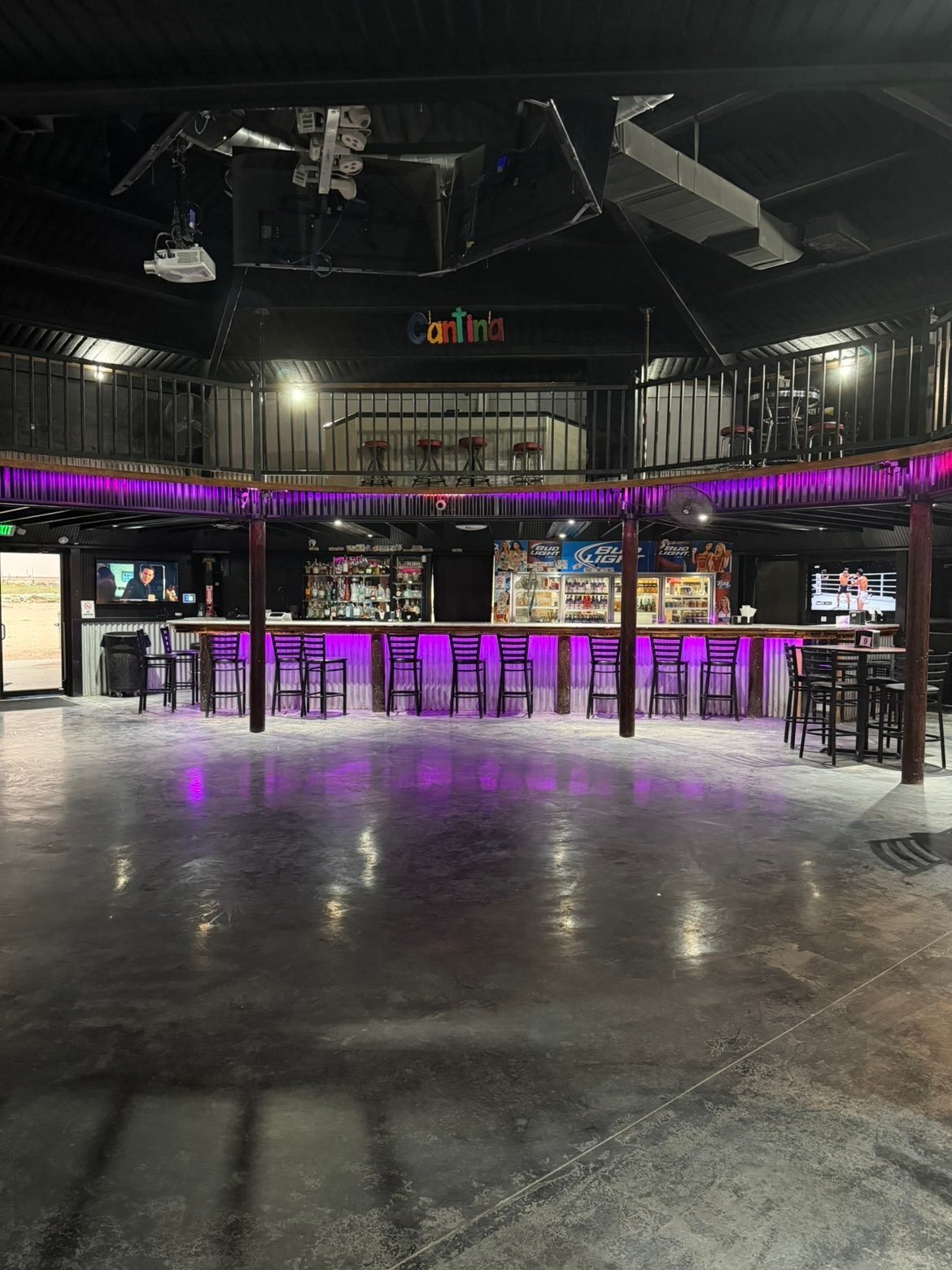 Modern nightclub interior with purple neon lighting, bar counter with stools, and dark concrete flooring under industrial ceiling