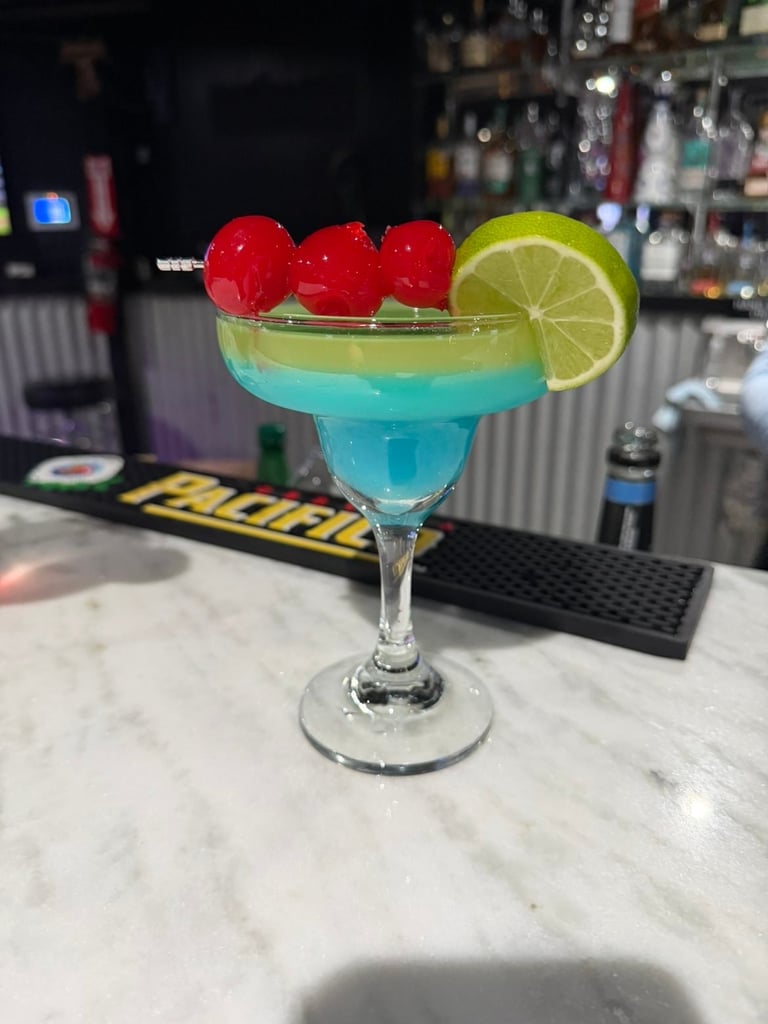 Blue cocktail in a margarita glass garnished with red maraschino cherries and lime wheel at a bar counter